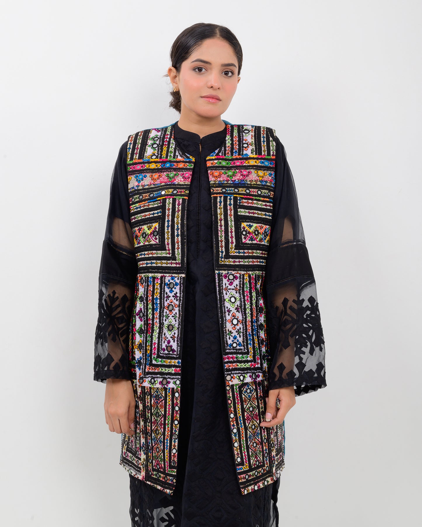 Woman wearing a colorful balochi embroidered vest over a black outfit on a white background