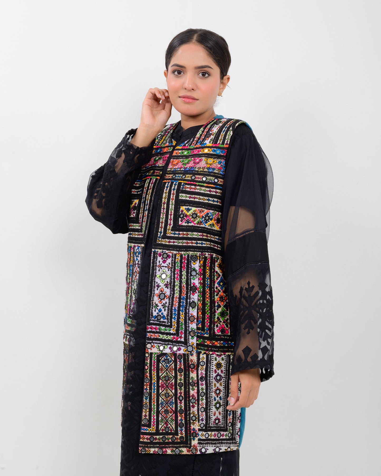 Woman wearing a colorful balochi embroidered vest over a black outfit on a white background
