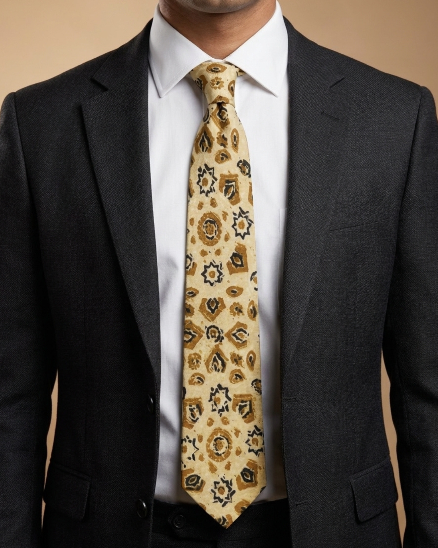 A man wearing a dark gray suit jacket, white dress shirt, and a patterned beige necktie with brown and black details.