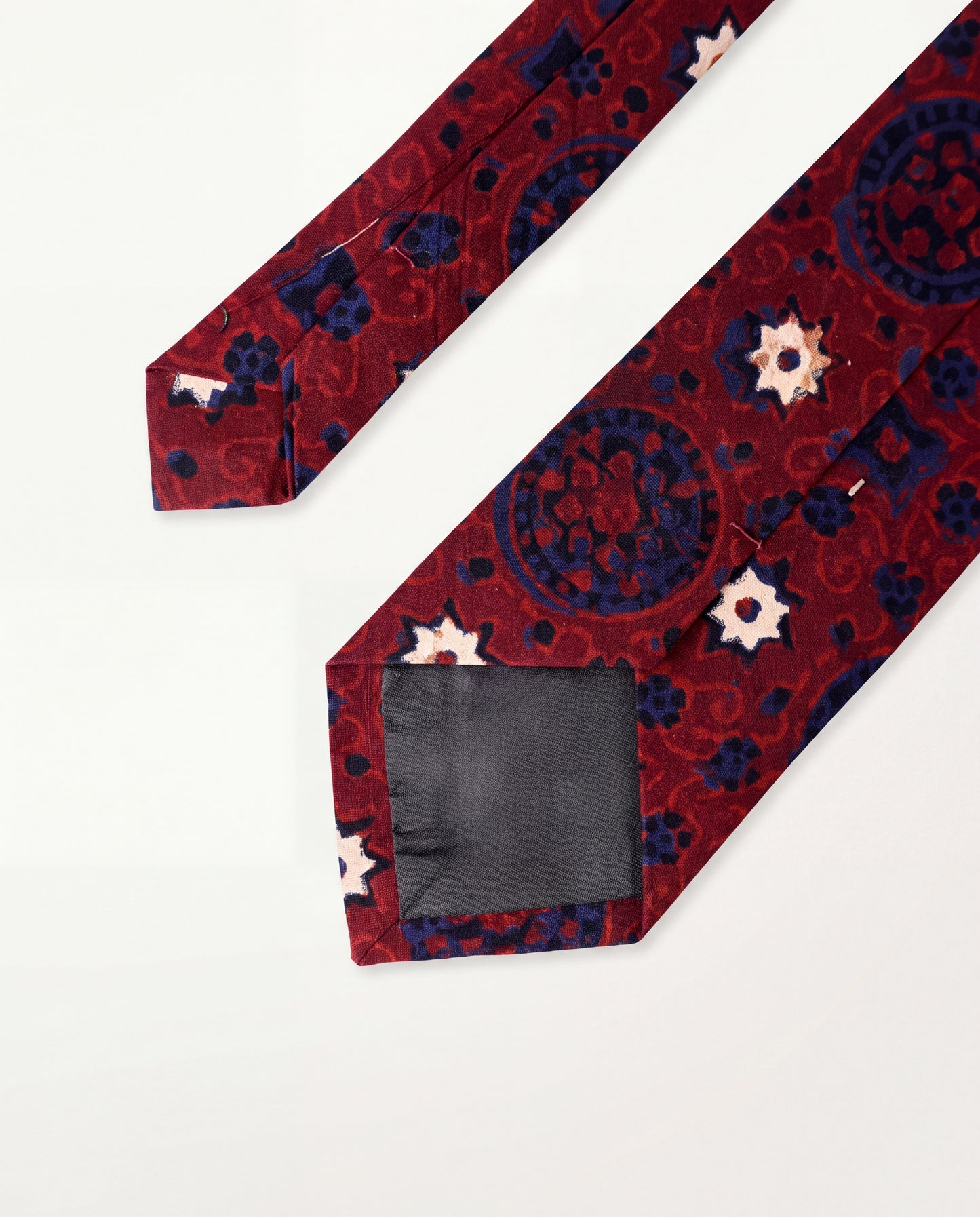 A patterned necktie with a deep red base, navy blue medallions, and small white and pink floral accents, displayed on a clean white background with the tip showing a dark gray lining.