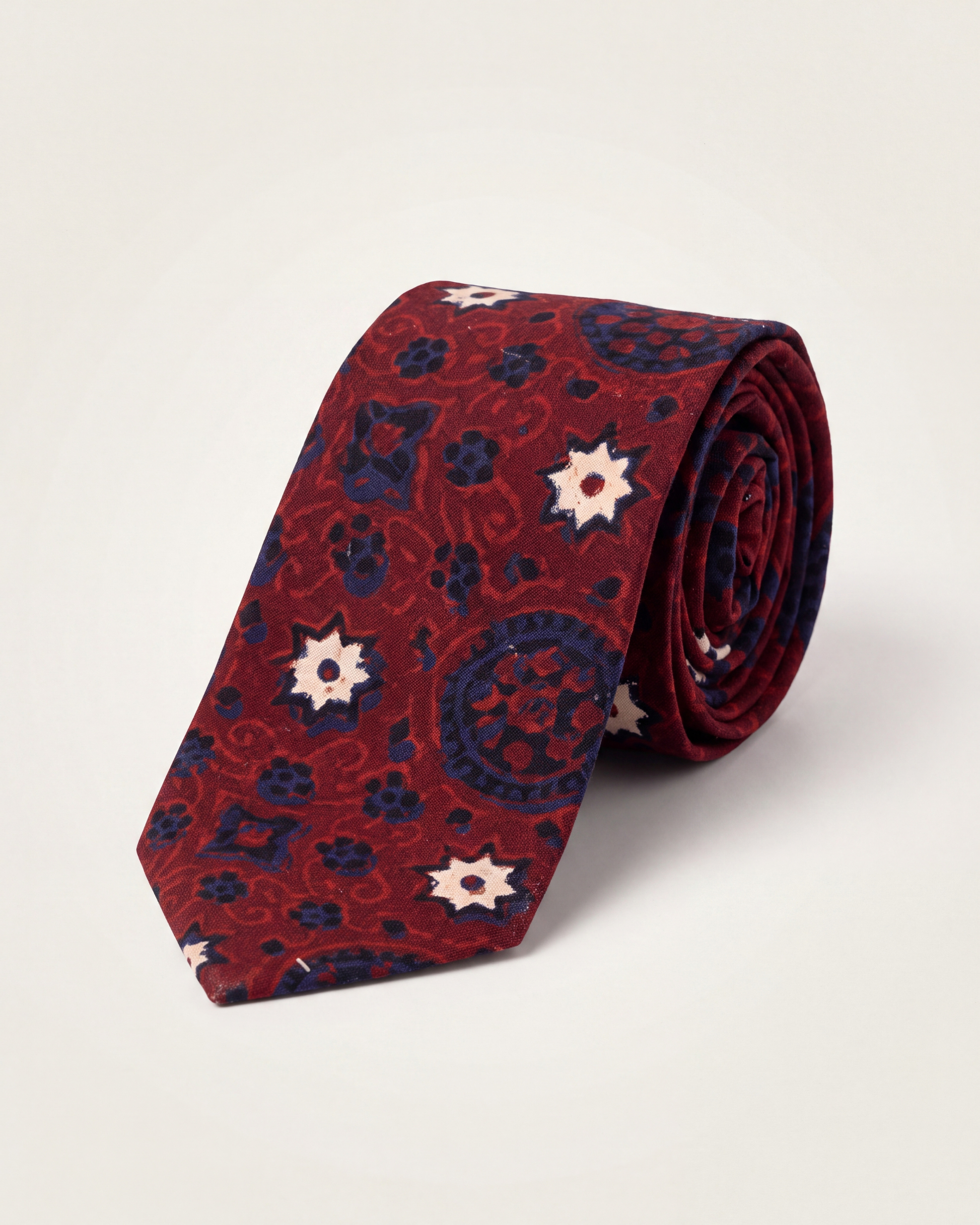 A rolled Cotton necktie featuring a red and navy blue paisley Ajrak pattern with cream starburst accents, displayed on a light beige background.