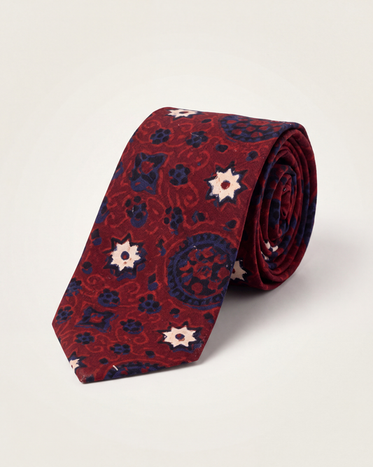 A rolled Cotton necktie featuring a red and navy blue paisley Ajrak pattern with cream starburst accents, displayed on a light beige background.