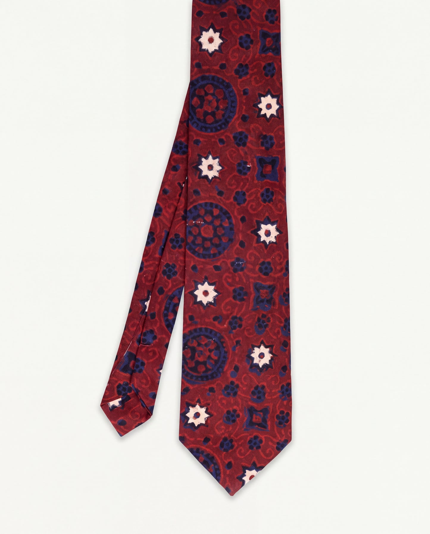 A patterned cotton tie with a deep red base, featuring navy blue circular medallions and cream-colored starburst motifs, displayed flat against a light off-white background.