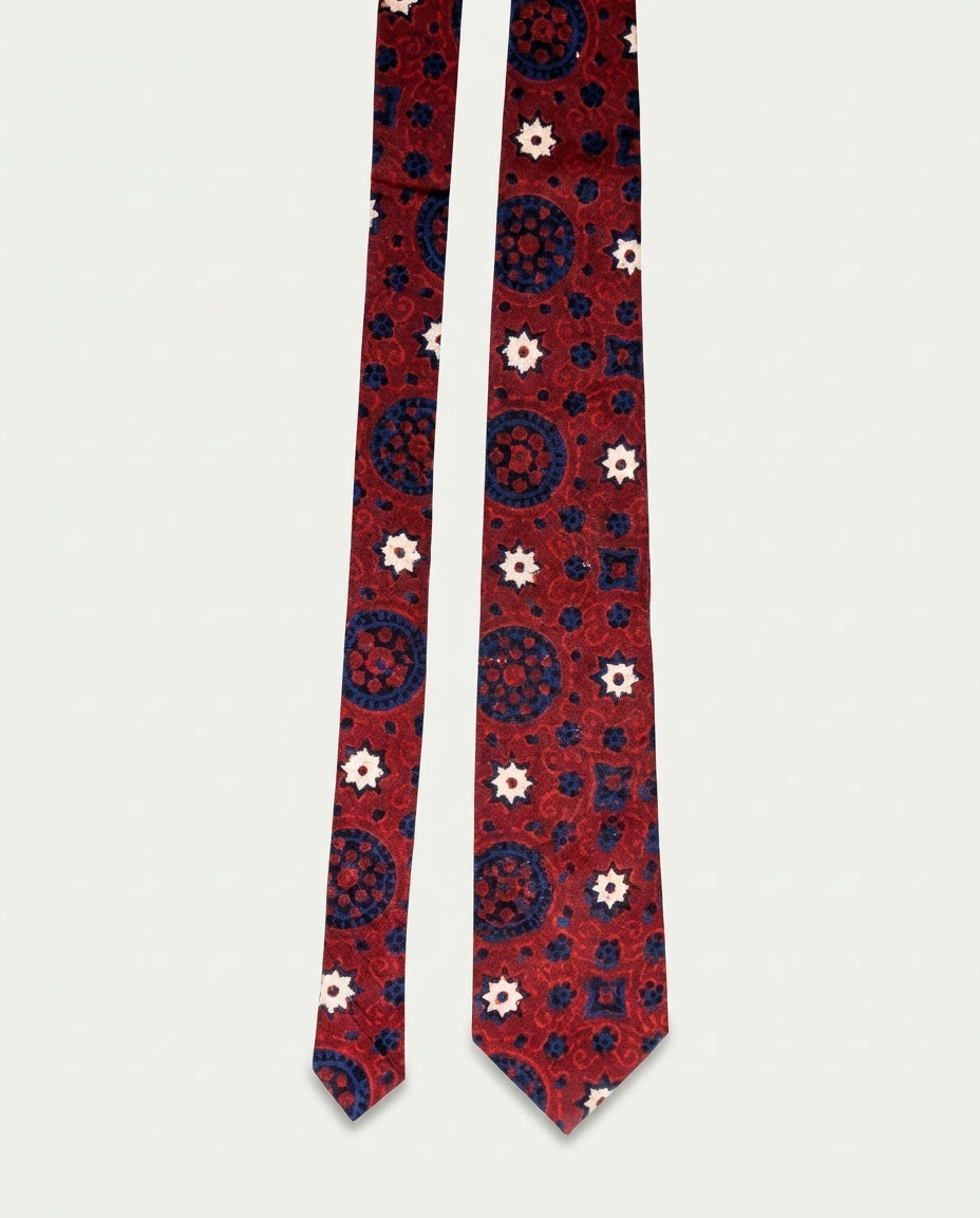 A red necktie with a detailed pattern of navy blue medallions and small white flowers, laid flat against a light gray background.
