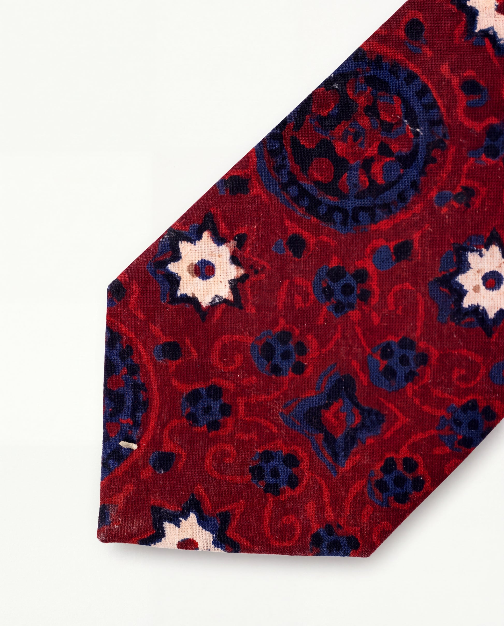 A close-up of a patterned necktie with a deep red base, navy blue floral and scroll motifs, and cream starburst accents, laid diagonally on a plain white background.