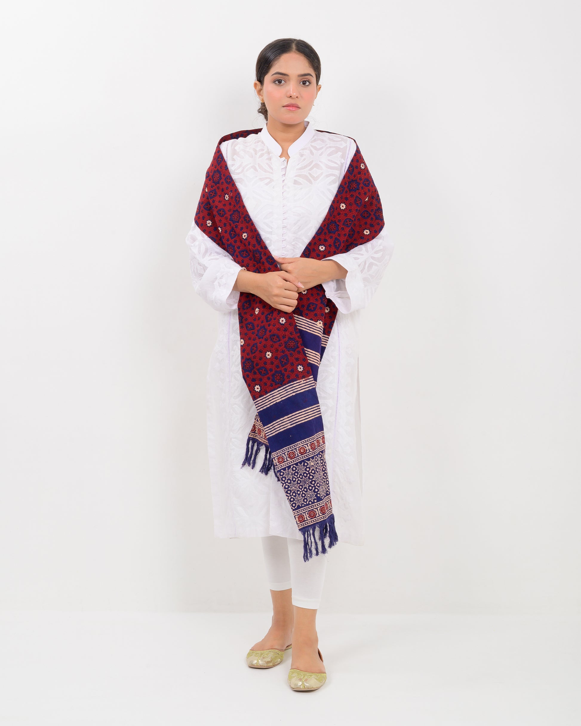 A woman wearing a white outfit with a patterned red and blue block print ajrak stole scarf on a white background