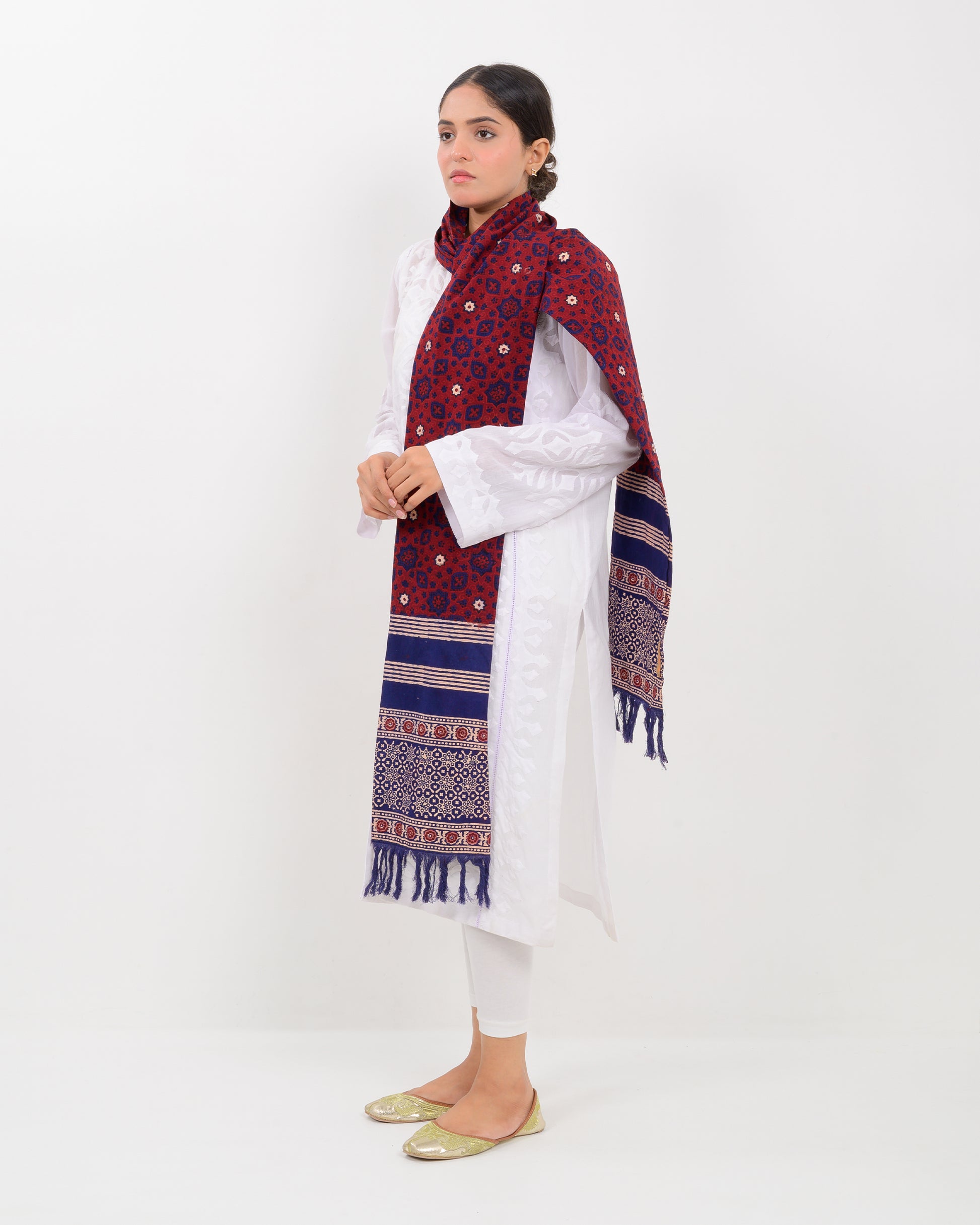 A woman wearing a white outfit with a patterned red and blue block print ajrak stole scarf on a white background