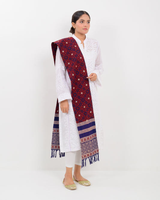 A woman wearing a white outfit with a patterned red and blue block print ajrak stole scarf on a white background