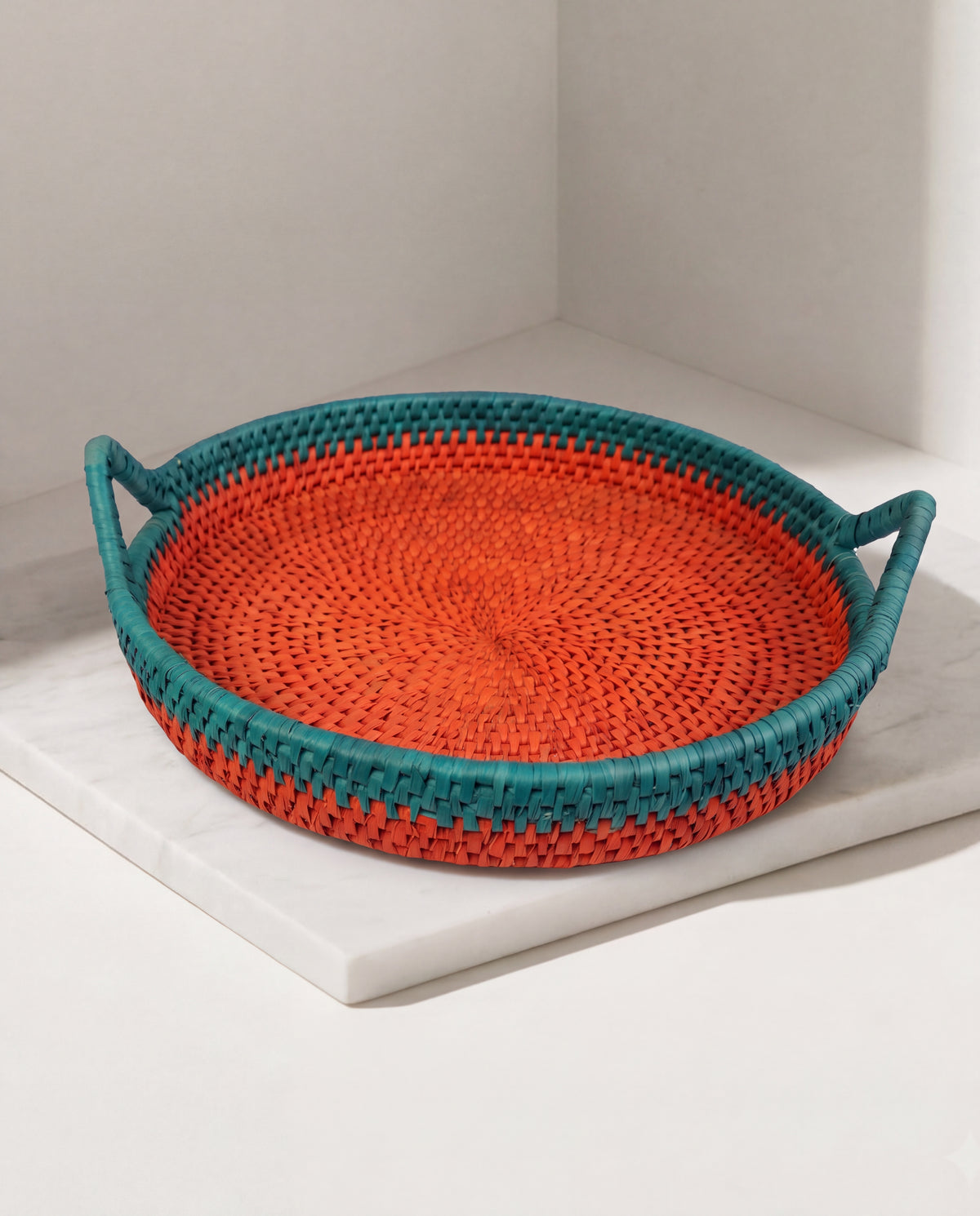 A round woven basket tray with two side handles, featuring a bright orange center and teal-blue outer rim, displayed on a white marble slab in a minimalist corner setting.