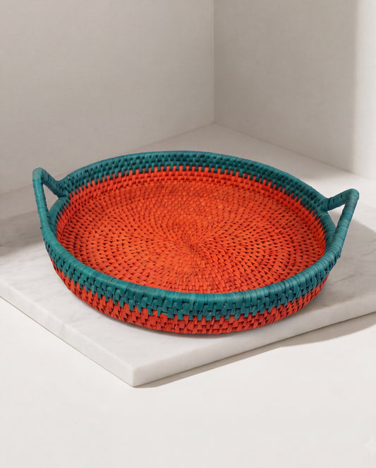 A round woven basket tray with two side handles, featuring a bright orange center and teal-blue outer rim, displayed on a white marble slab in a minimalist corner setting.