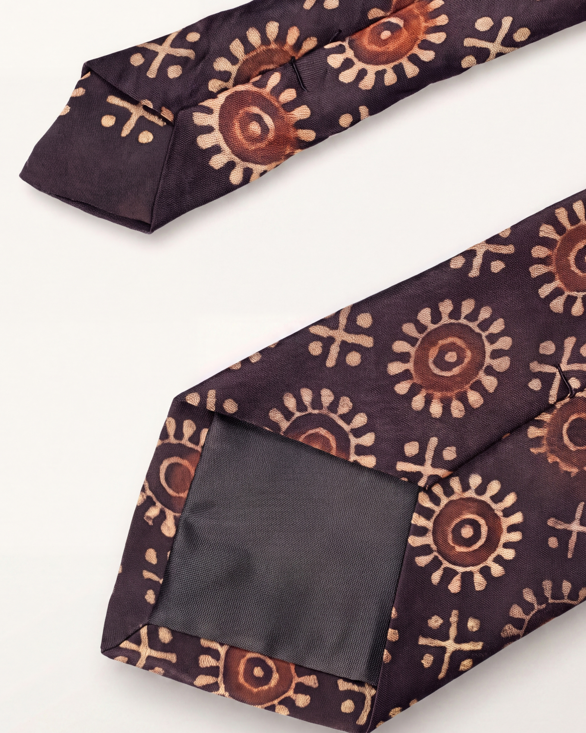 A dark purple tie with a brown and beige block-printed circular sunburst pattern, shown folded on a white background.