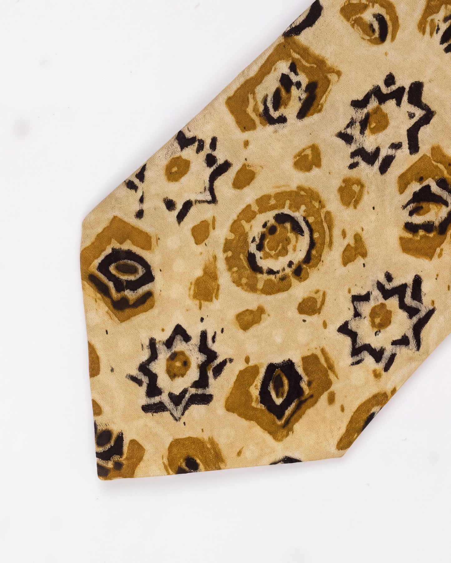 A close-up of a necktie with a beige base and a batik-style pattern in brown, black, and white, shown on a plain white background.