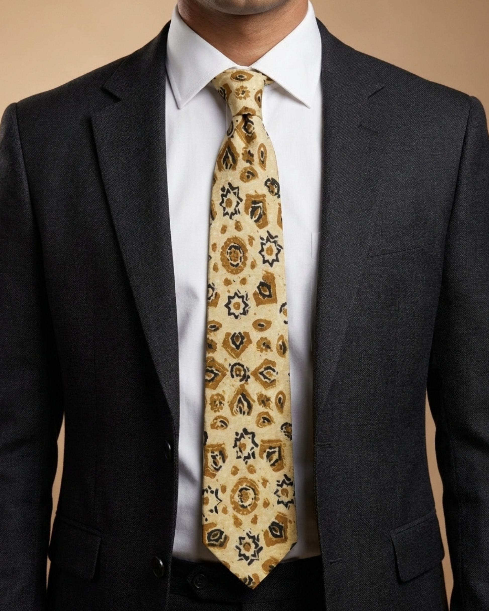 A man wearing a dark gray suit jacket, white dress shirt, and a patterned beige necktie with brown and black details.