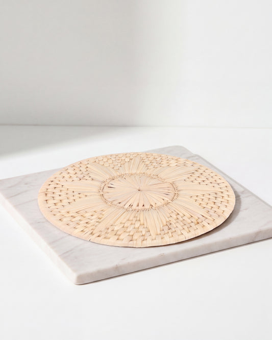 A round woven natural-fiber placemat with a sunburst pattern, displayed on a square white marble board against a clean white background.
