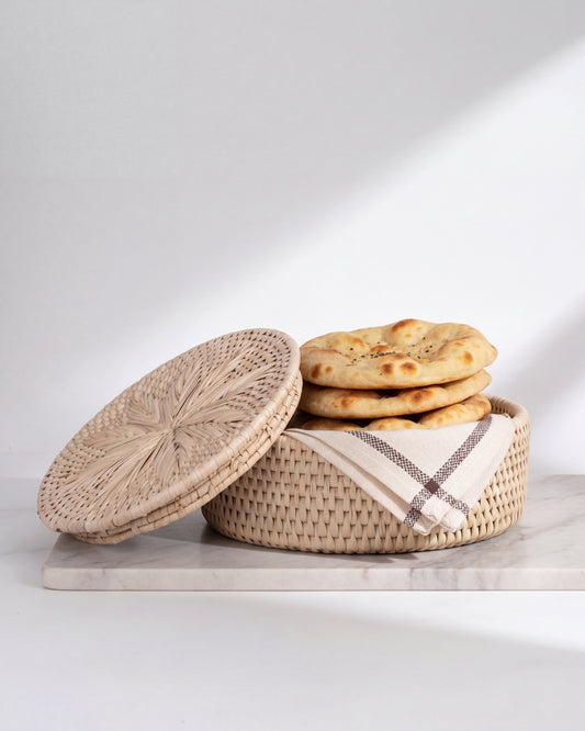 A round woven basket with a matching lid, shown on a light stone surface against a white background. The basket is filled with flatbread and a folded cream cloth with a brown stripe.