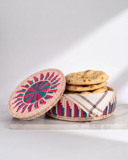 A round woven basket with a matching lid, styled with flatbread and a striped cloth, set on a light marble board against a white background with soft shadows.