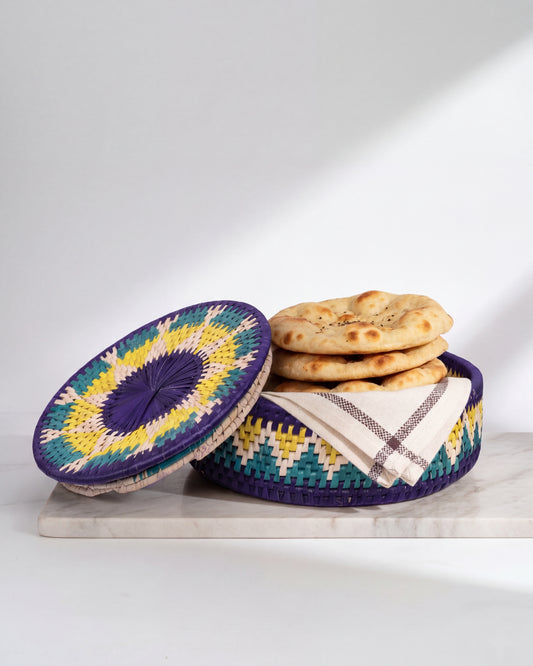 A round woven basket with a matching lid, decorated with a purple, yellow, and teal geometric pattern, sits on a marble board. Inside the basket is a stack of flatbread and a folded cream-colored checkered cloth.