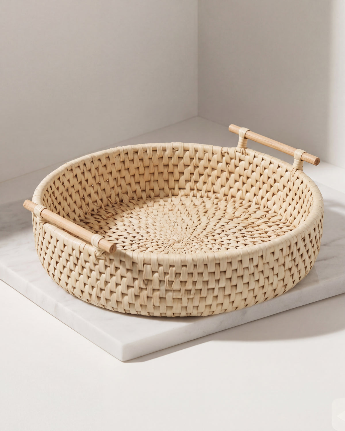 Round woven natural rattan tray with two light wood handles, shown on a white marble slab against a neutral light gray background.