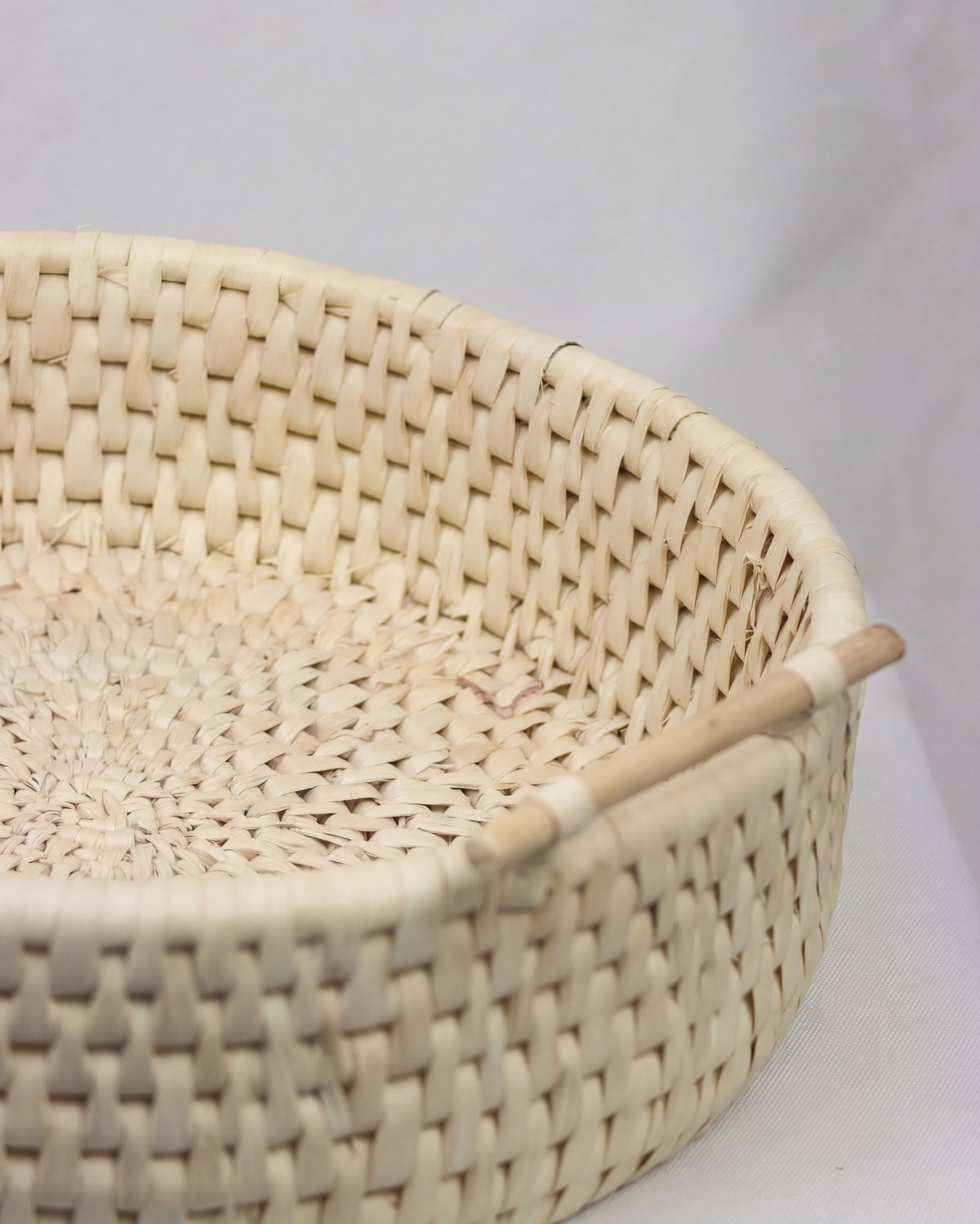 A close-up of a round woven basket made from natural light beige fibers, featuring a simple wooden handle on a light gray background.
