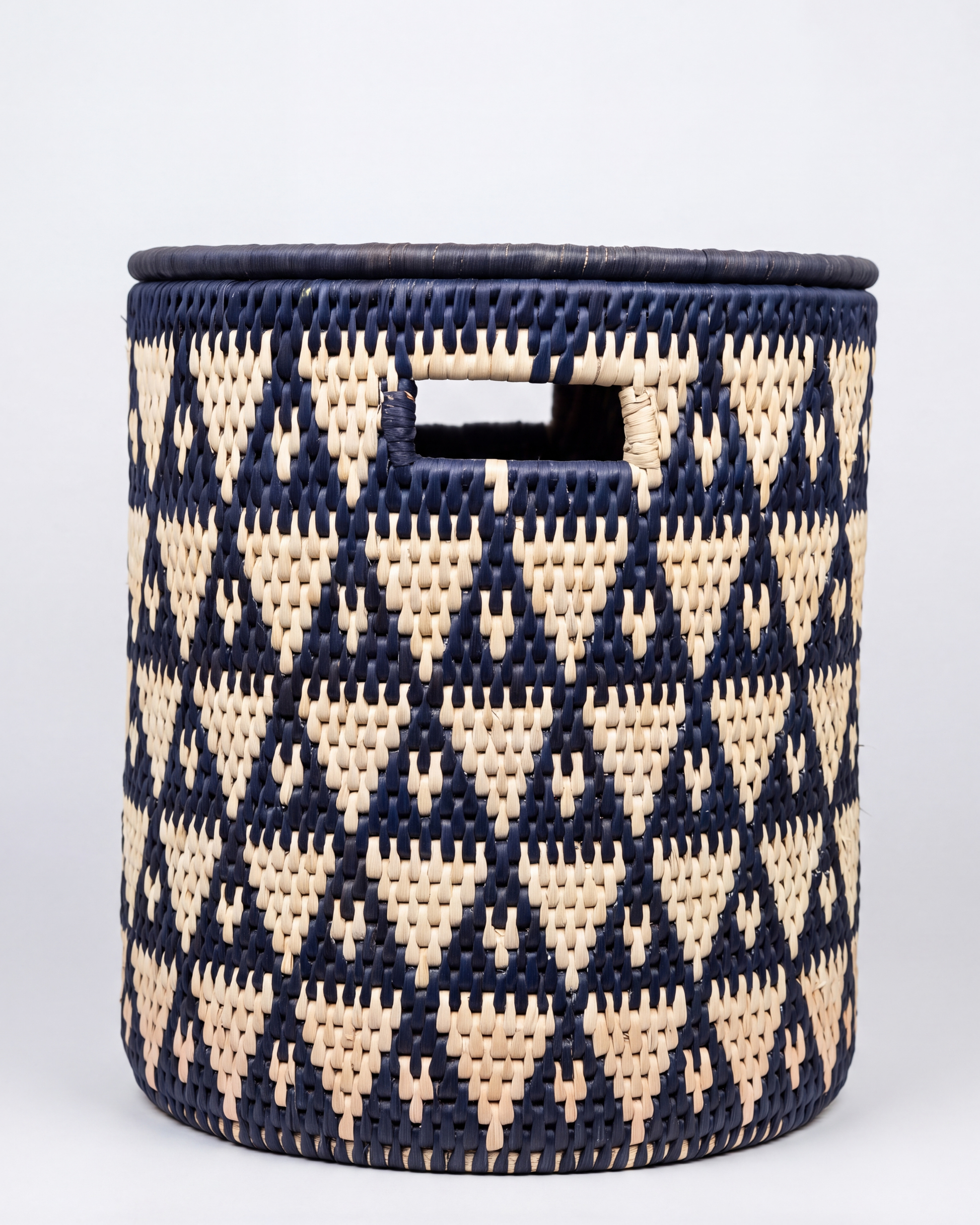 A round woven basket with a navy blue and natural beige geometric pattern, featuring a cutout handle on the front, set against a plain light gray background.