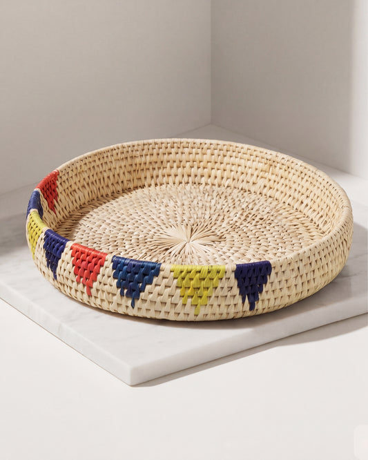 A round woven tray made of natural straw with colorful geometric patterns in red, blue, yellow, and purple around the rim, displayed on a white marble slab against a light gray background.